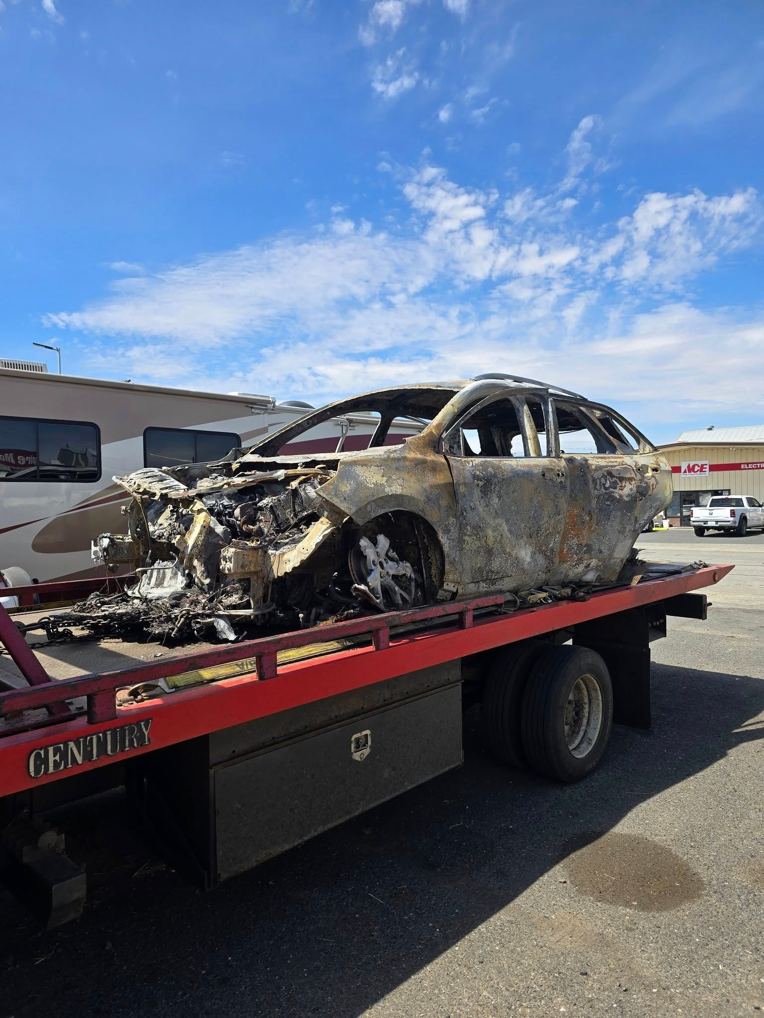Burnt-out car on a flatbed tow truck. Remains of vehicle are charred. Bright blue sky overhead.