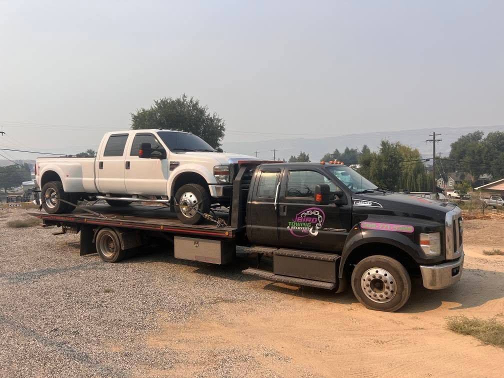A black tow truck carries a white pickup truck on a gravel lot under a hazy sky.