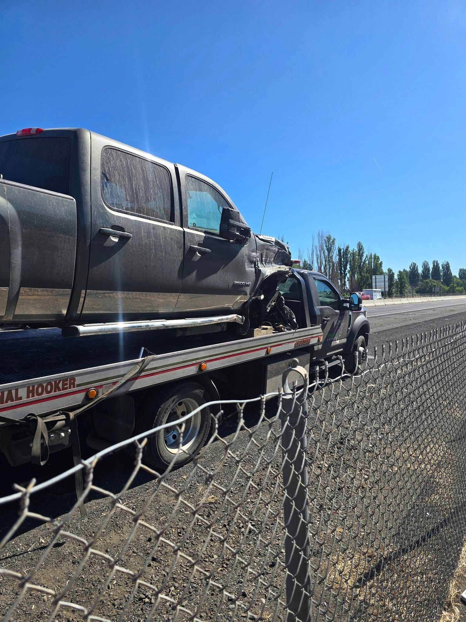 Gray pickup truck loaded onto a flatbed tow truck next to a chain-link fence on a sunny day.