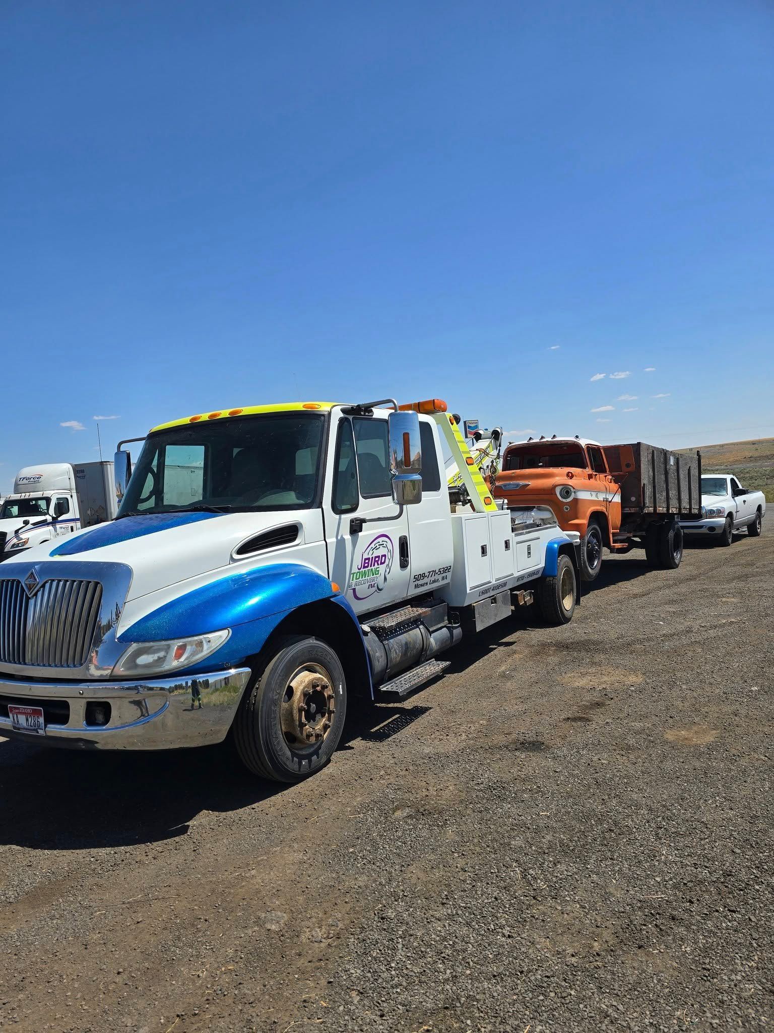 Tow truck towing an orange truck and a trailer on a gravel road under a blue sky.