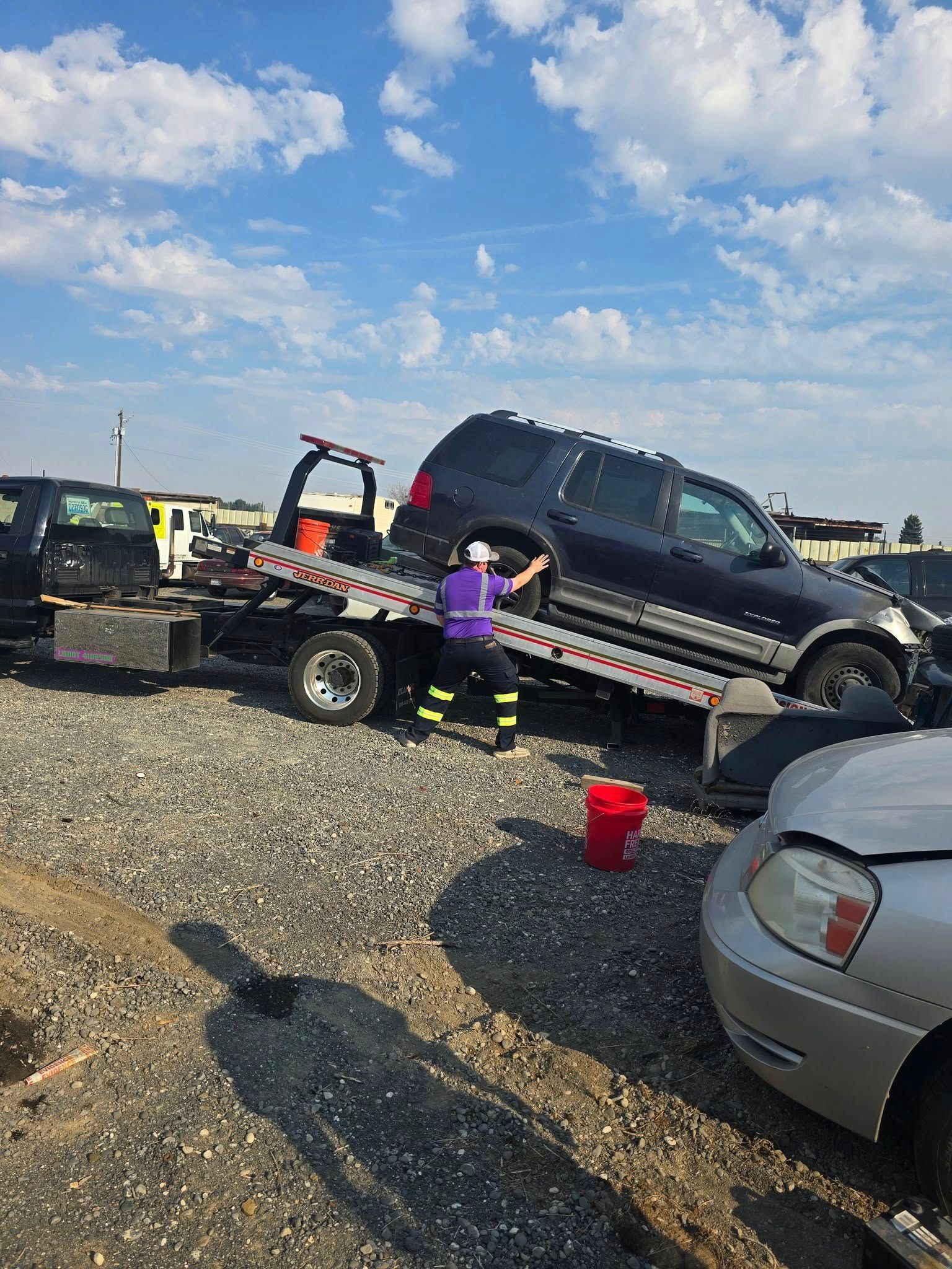 Tow truck loading a dark SUV in a junkyard on a sunny day. A person in safety gear assists.
