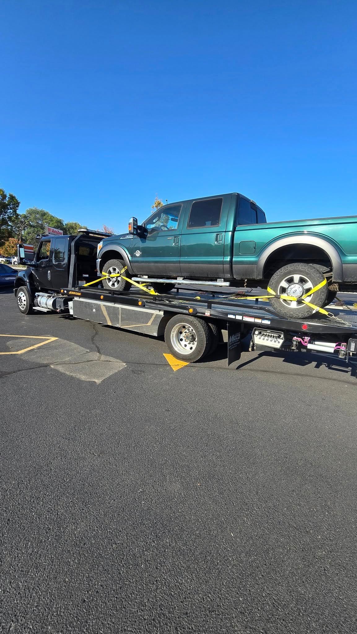 Green pickup truck secured on a flatbed tow truck in a parking lot on a clear day.