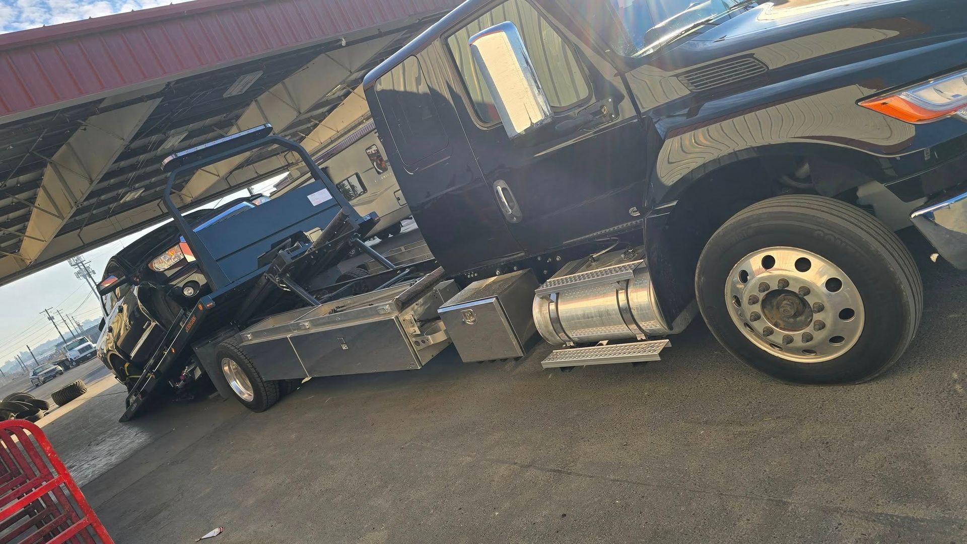 Black tow truck carrying a blue vehicle on its flatbed, parked under a red awning.
