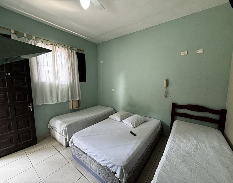 Um quarto de hotel com paredes verde-claras apresenta uma cama de casal ladeada por duas camas de solteiro e uma porta à esquerda.