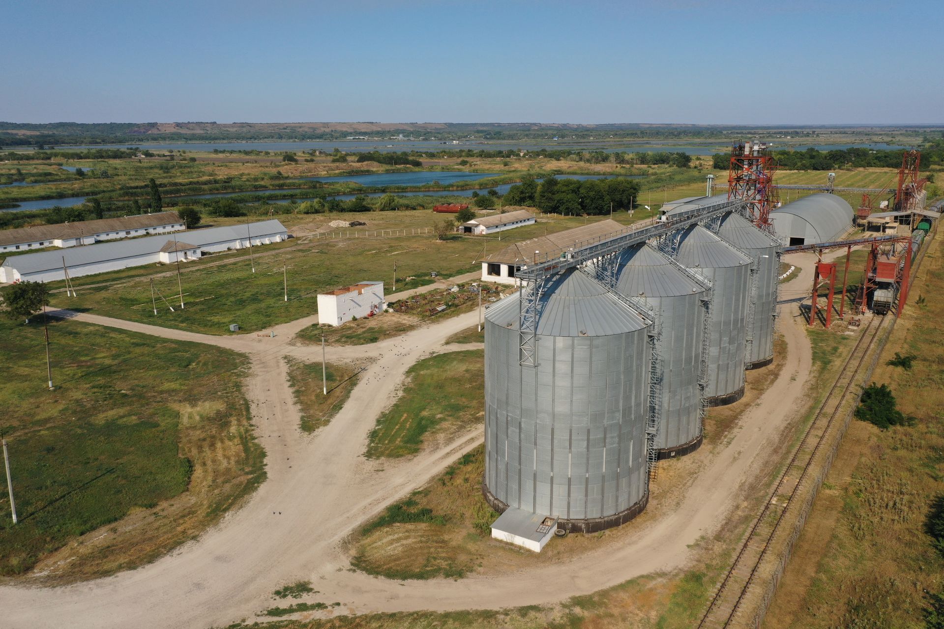 Aerial view of a grain storage facility with tall silos, buildings, and surrounding fields under a blue sky.