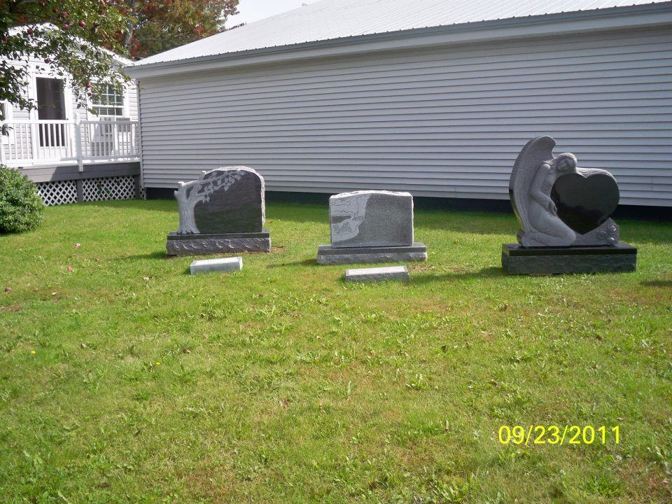 Three gravestones in a grassy yard; one with a tree, one plain, one with angel and heart.