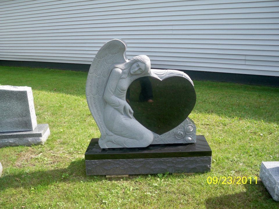 Granite angel kneeling, embracing a heart-shaped black granite memorial. Outdoors on grass.