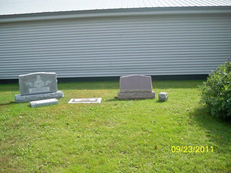 Graveyard scene with several headstones on a grassy lawn in front of a white building; date stamp 