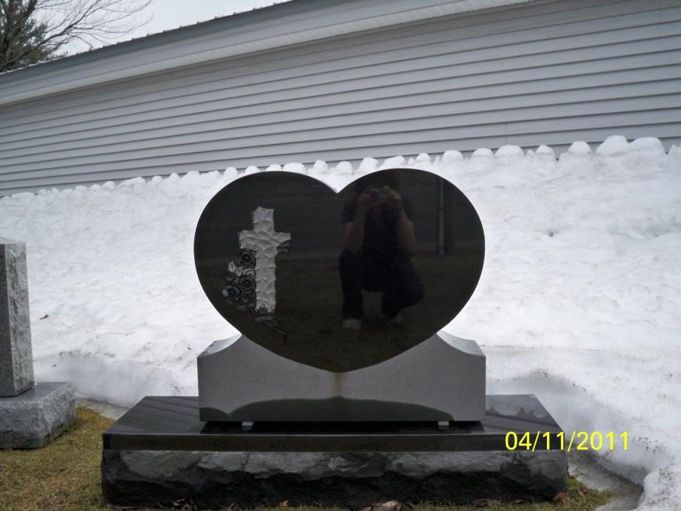 Heart-shaped black tombstone with a cross, reflecting a person taking a photo, set in a snowy area.