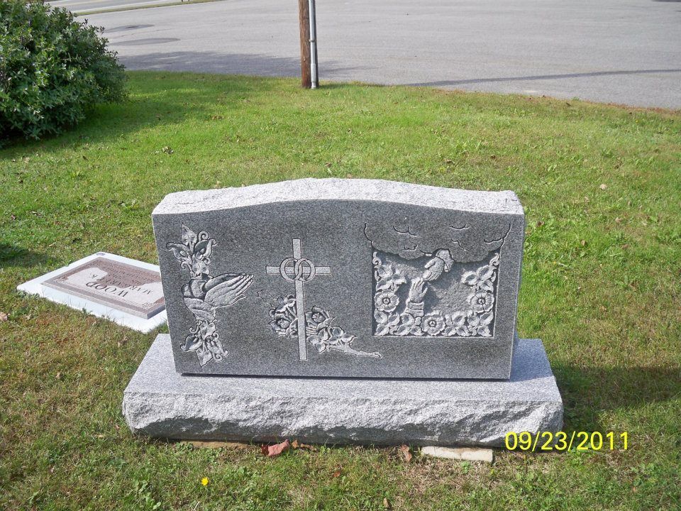 A gray headstone with etched images: a rooster, cross, and a scene of Jesus, in a grassy setting.