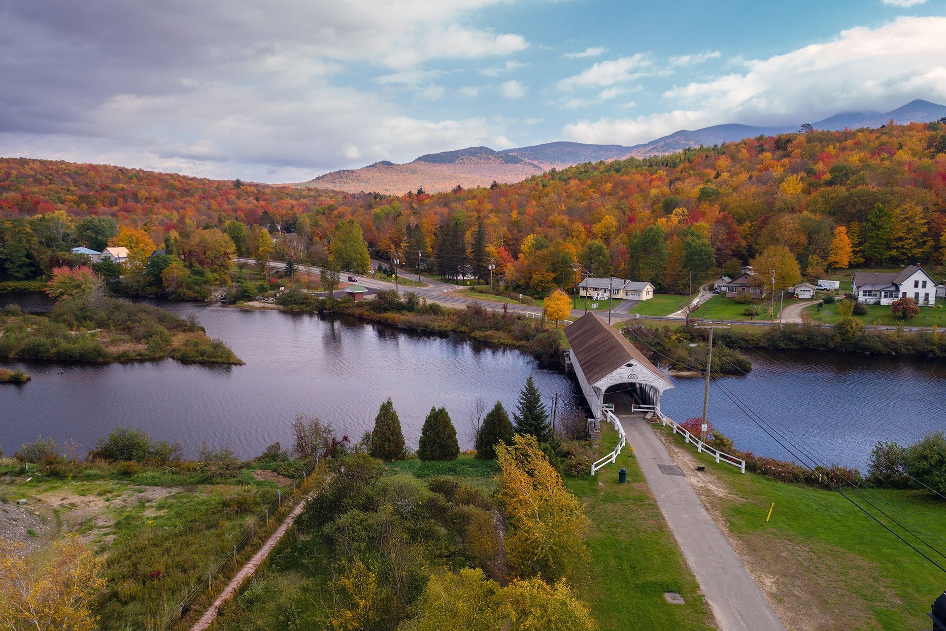 Covered bridge over a river in autumn; colorful foliage, mountains in the background.