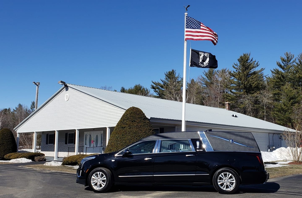 Black hearse parked outside a funeral home under US and POW flags. Clear blue sky.