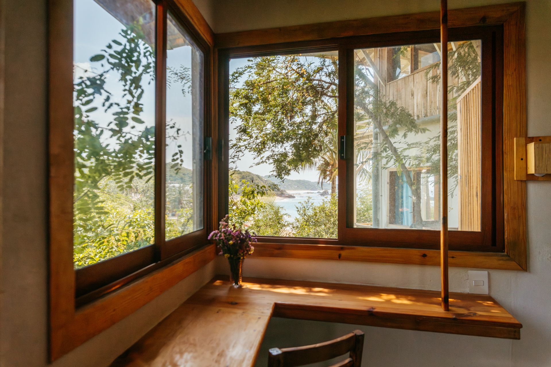 Ventana esquinera con marco de madera que da a un paisaje exuberante y al agua en posada del arquitecto en mazunte, oaxaca