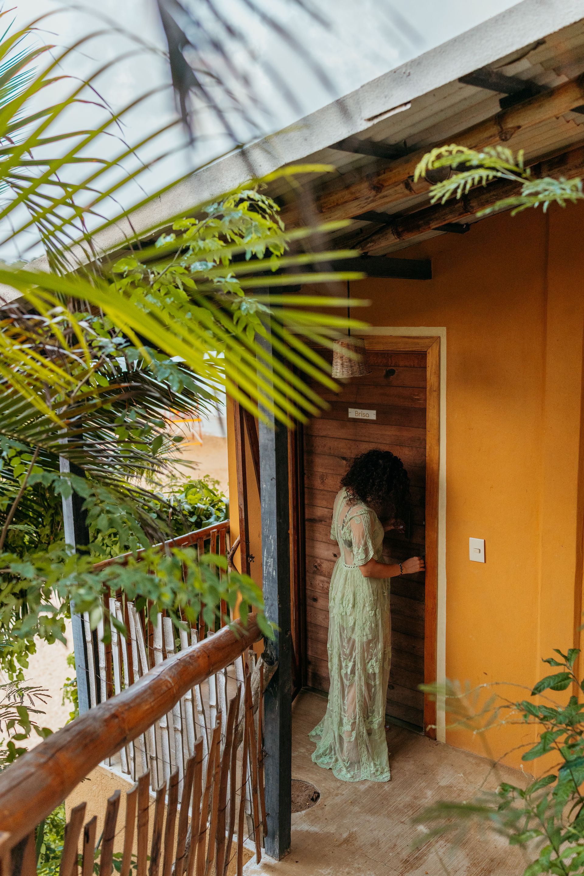 Mujer con vestido verde cerca de la puerta, balcón con barandilla de madera, pared amarilla, plantas en posada del arquitecto