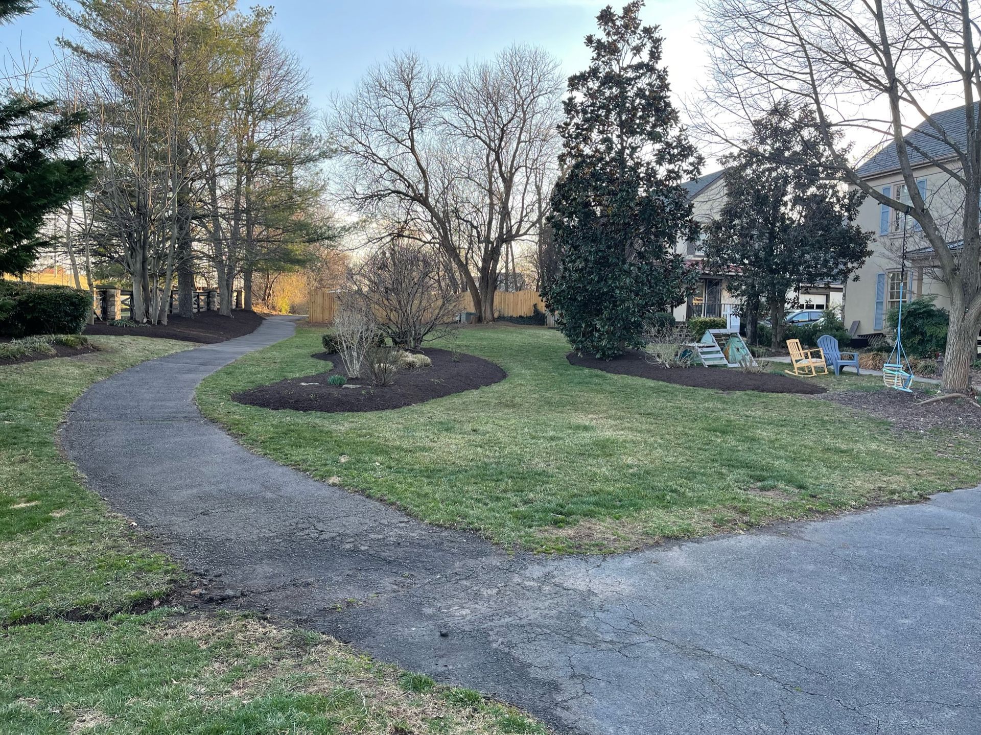 A path going through a lush green park with trees and a house in the background.