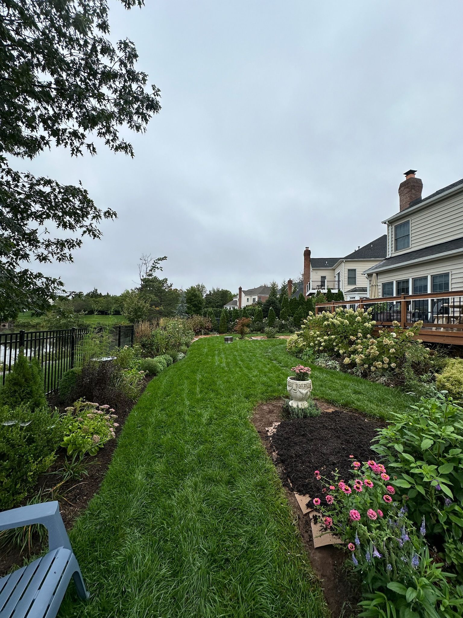 Lush green lawn path through a flower garden, leading to houses under an overcast sky.