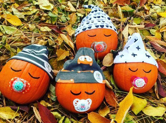 Four pumpkins wearing hats and pacifiers are sitting on a pile of leaves.