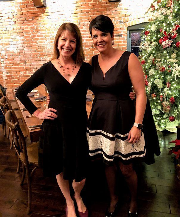 Two women in black dresses are posing for a picture in front of a christmas tree.