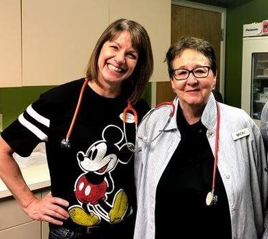 Two women are posing for a picture in a pediatrician's office . one of the women is wearing a mickey mouse shirt.