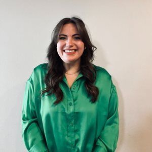 A woman in a green shirt is smiling and standing in front of a white wall.