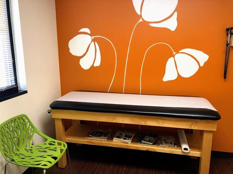 A green chair sits next to a wooden table in front of an orange wall with flowers painted on it