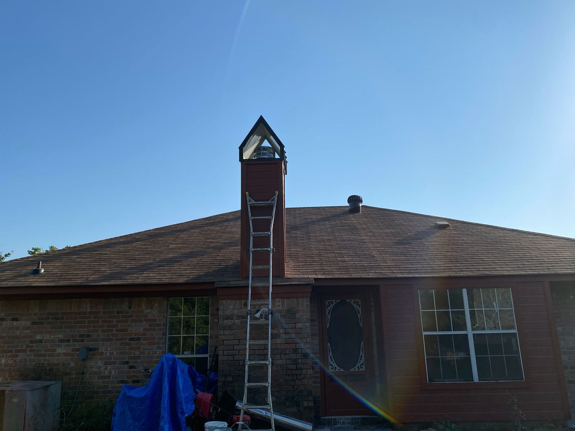 Ladder leaning against chimney on a house with a brown roof, blue tarp visible.