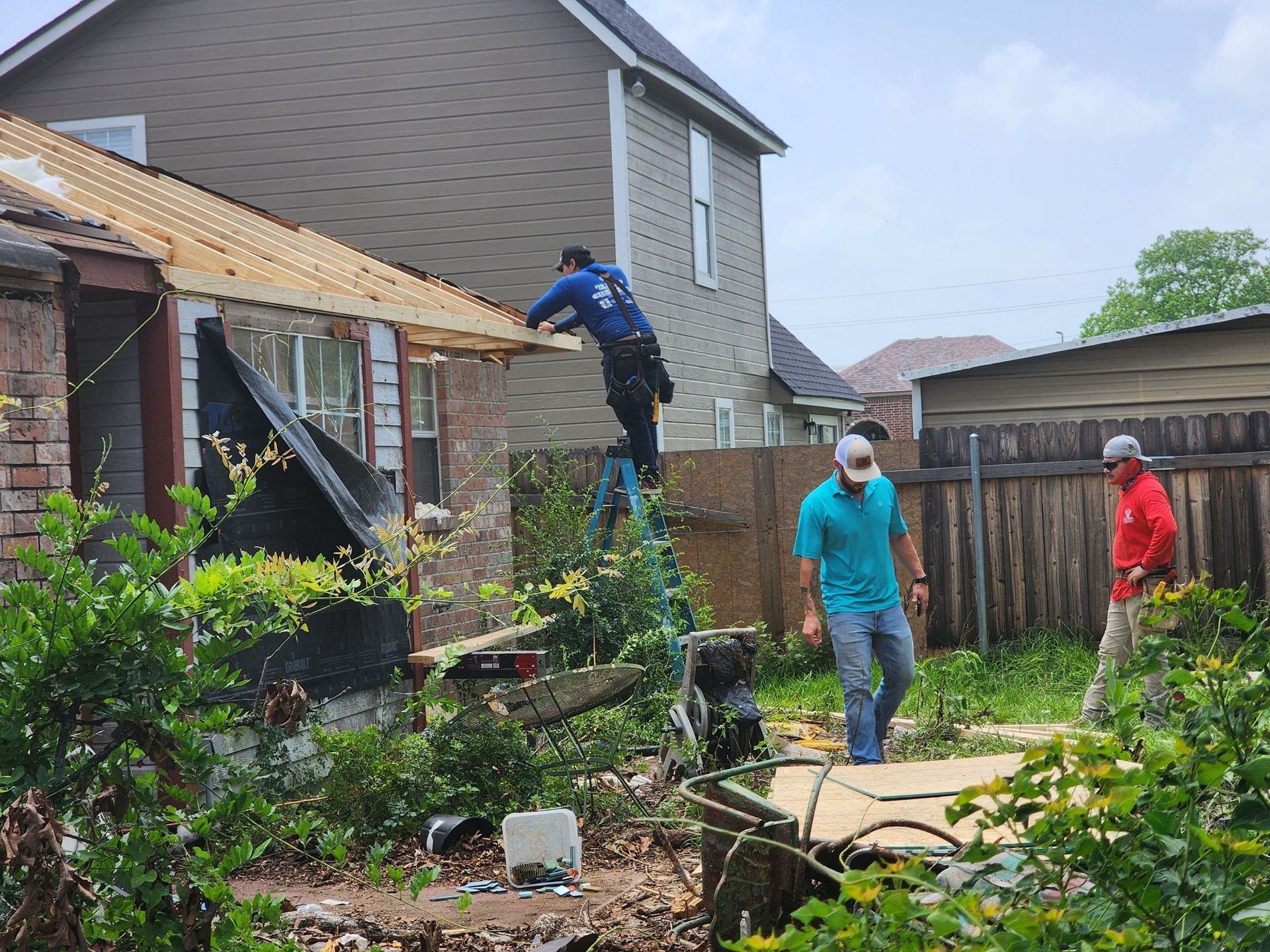 Three men working on a damaged house. One on a ladder, two others walking. Green foliage surrounds.