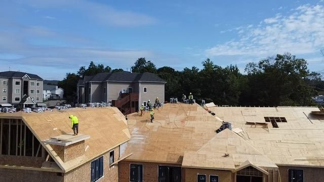 Construction workers on a roof, blue sky overhead. Buildings and trees in the background.