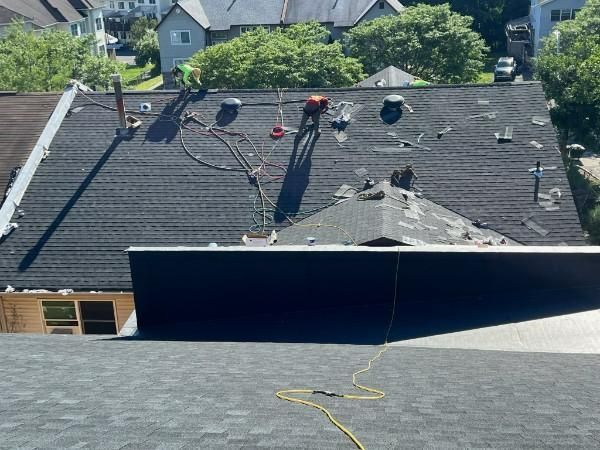 Workers on a dark-shingled roof, viewed from above, with tools and hoses. Green trees and buildings in the background.