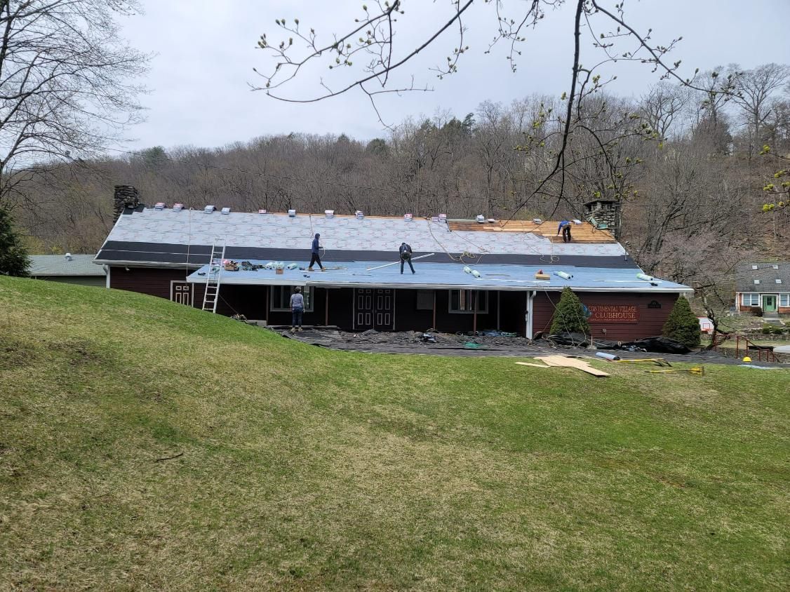 House with roof partially covered with new shingles, standing on a grassy hill, forest in the background.