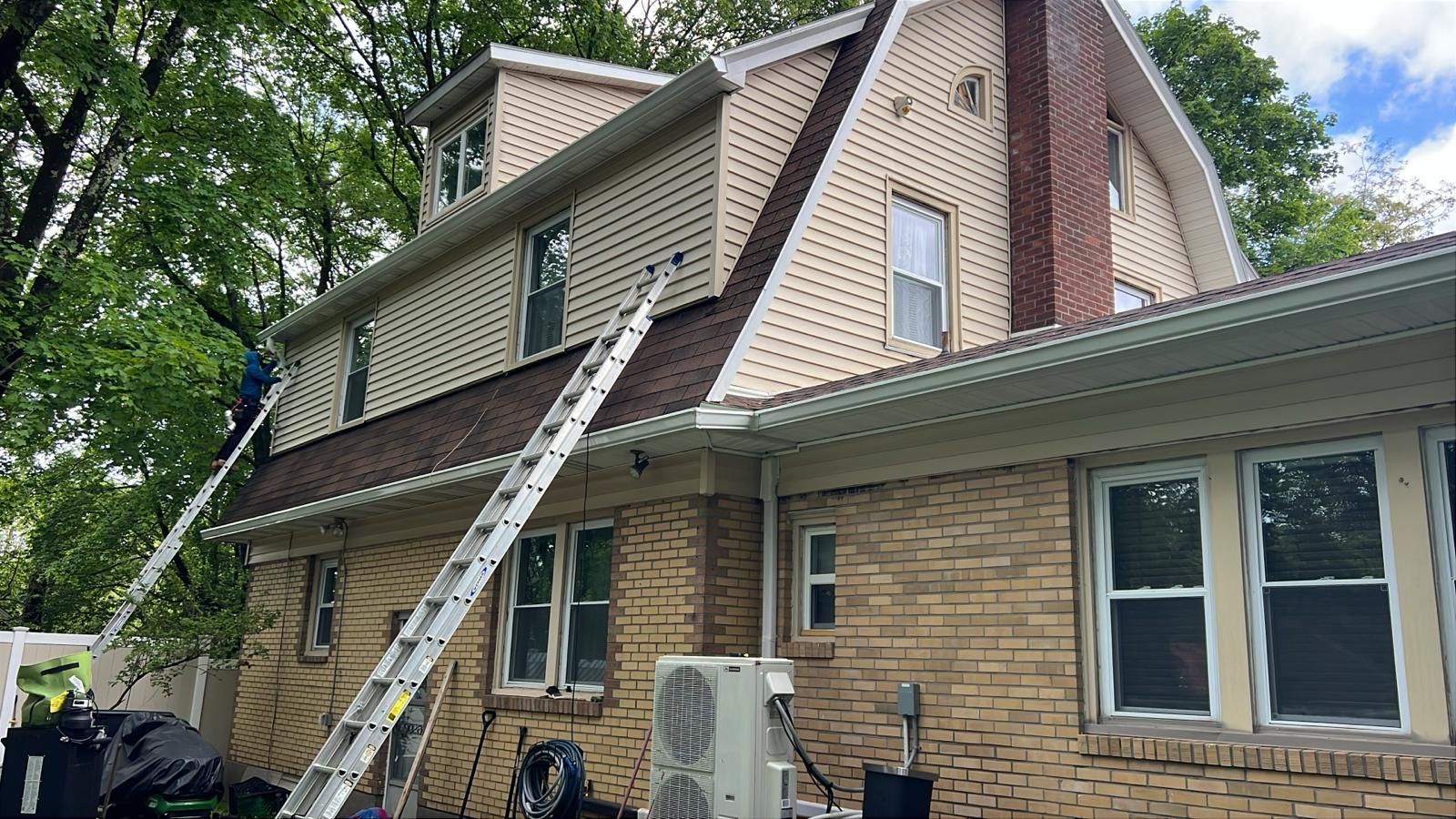 Two ladders on a brick house with a beige and brown roof; a person is working on the roof.