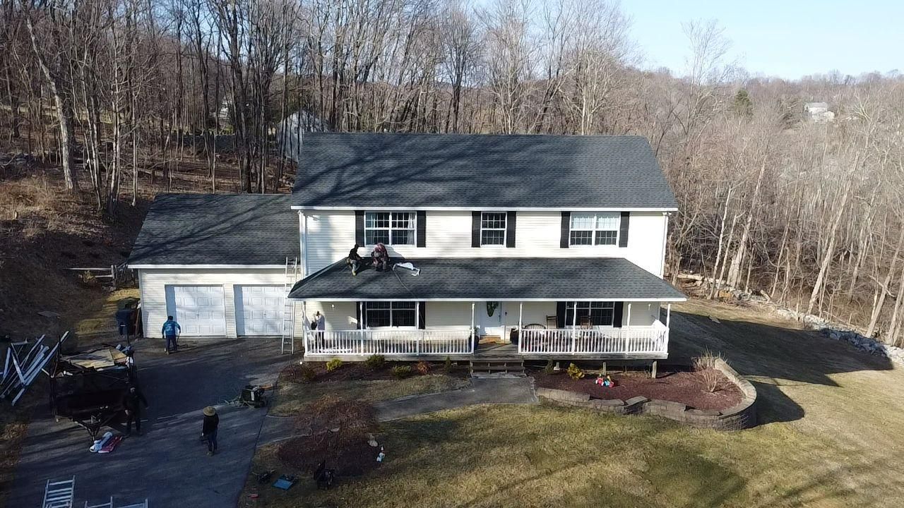 Two-story house with a porch, garage, and people on the roof, set in a wooded area.