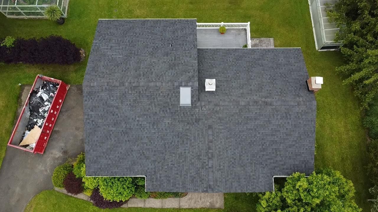 Overhead view of a dark gray roof on a house with a dumpster filled with debris next to it, surrounded by green grass and foliage.