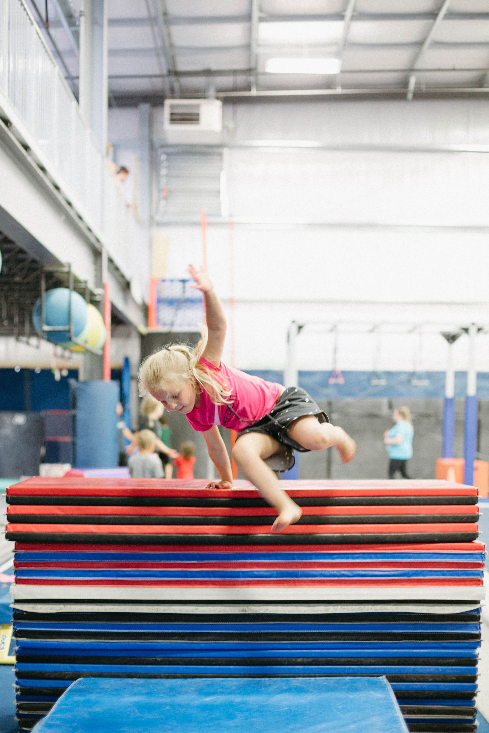 A little girl is jumping over a stack of mats in a gym.