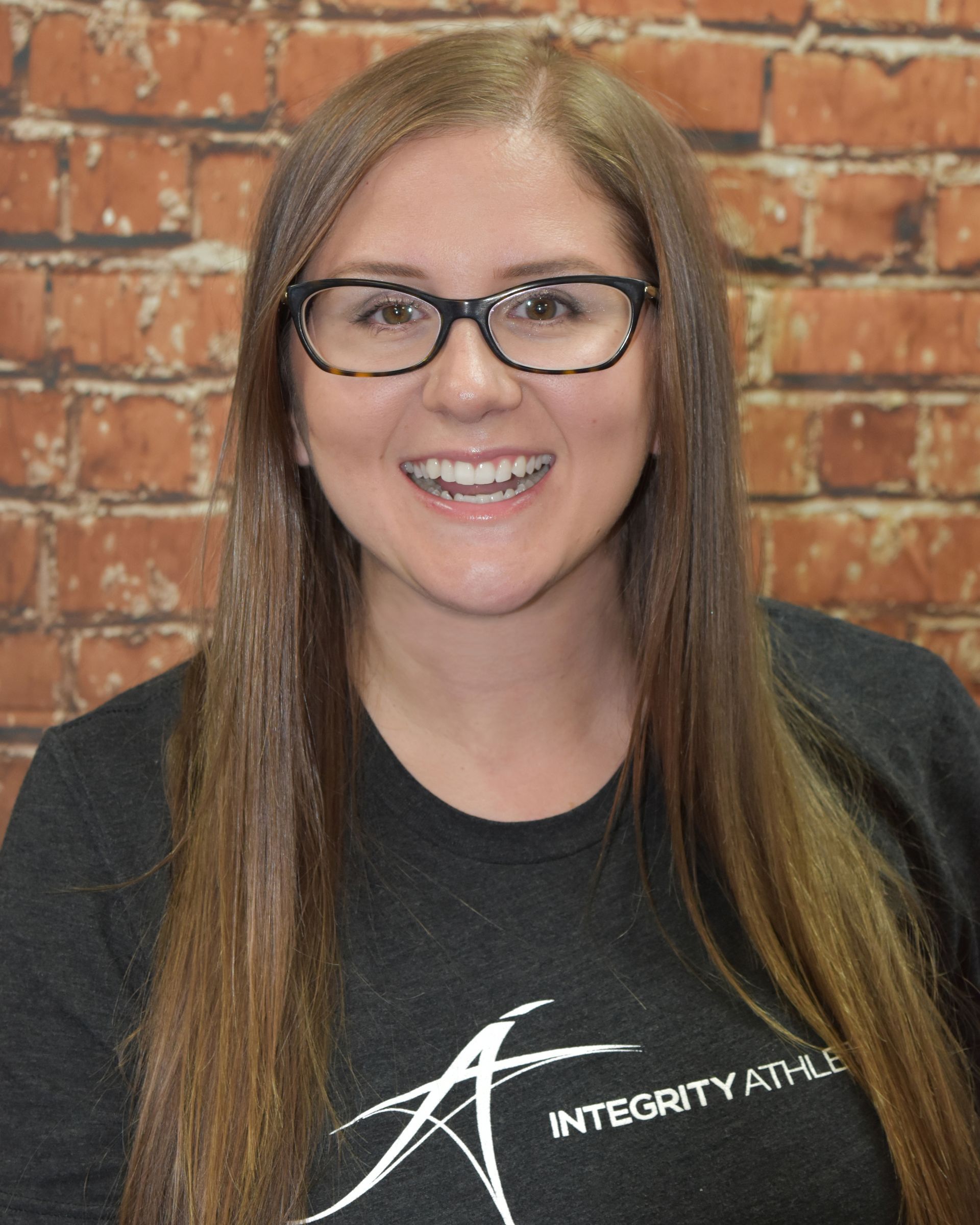 A woman wearing glasses and a black shirt with integrity written on it is smiling in front of a brick wall.