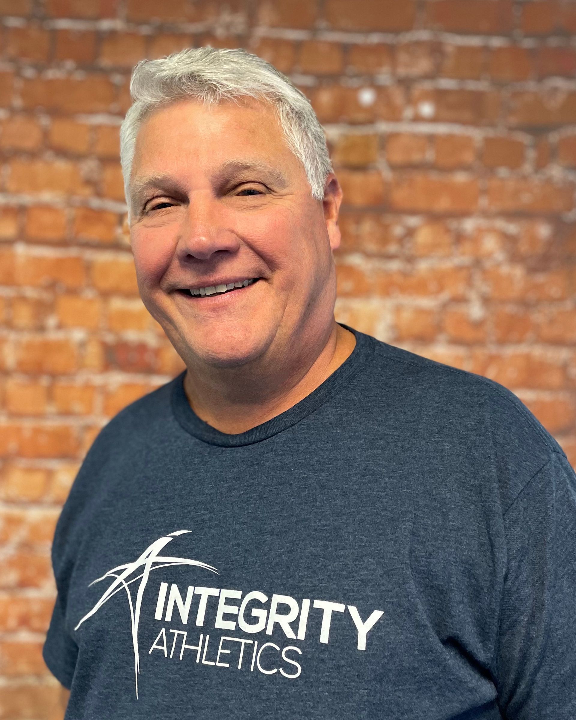 A man wearing a blue integrity athletics shirt is smiling in front of a brick wall.