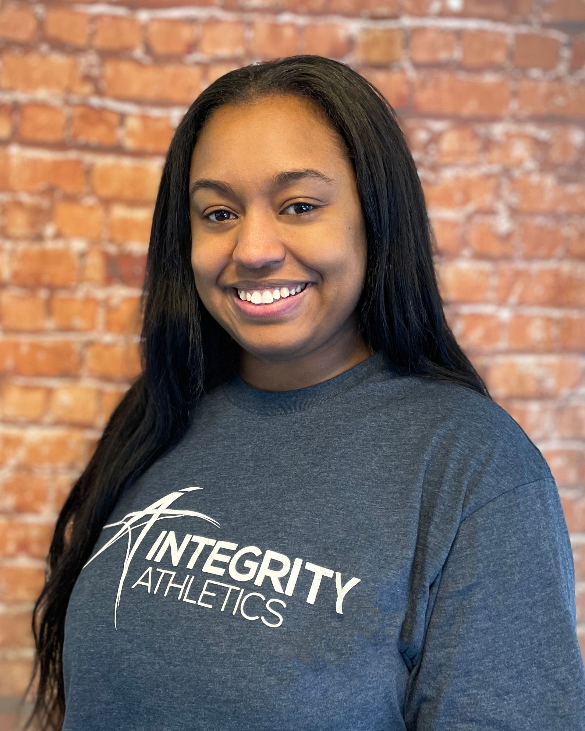 A woman wearing a integrity athletics shirt is smiling in front of a brick wall.