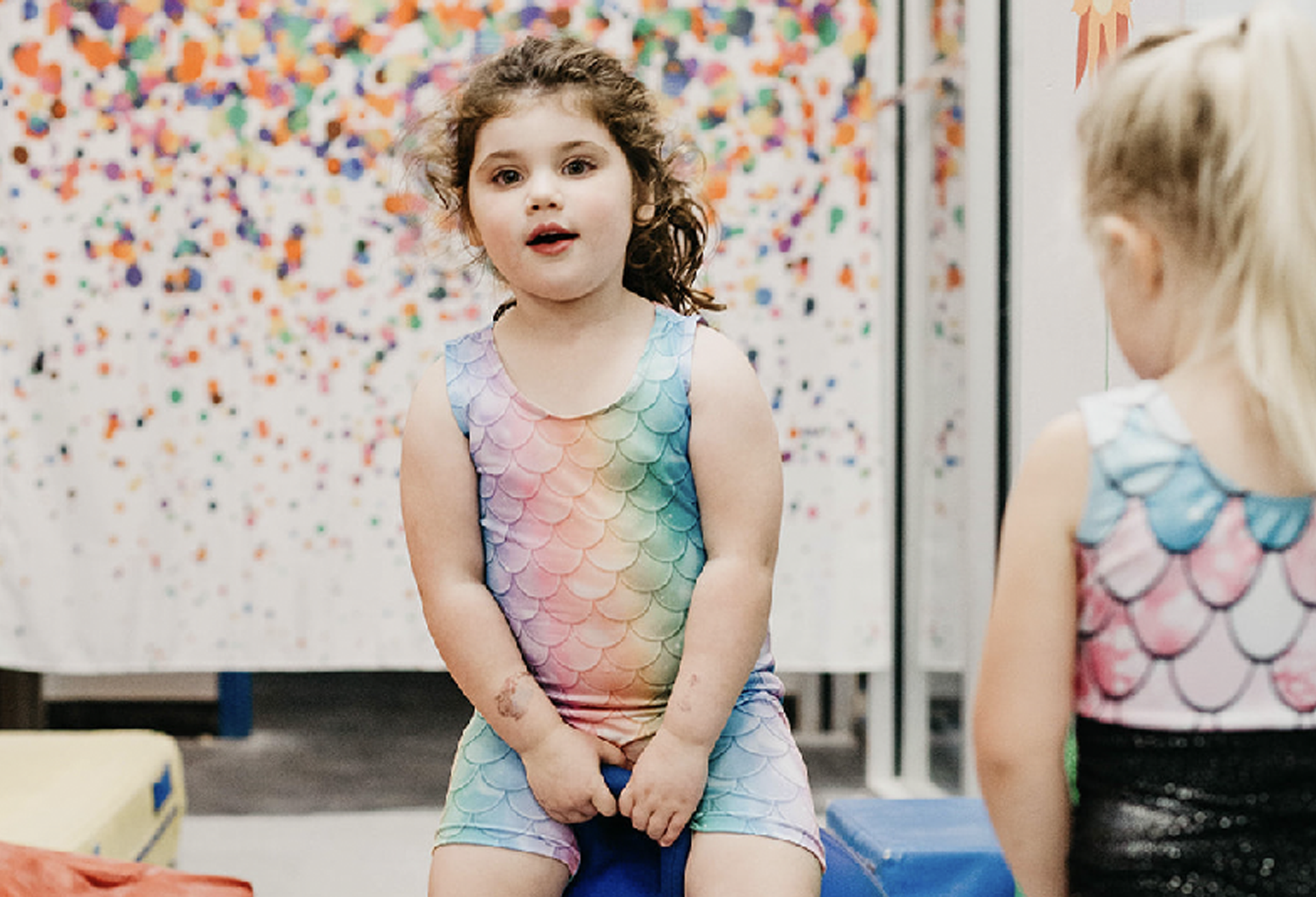 A little girl in a rainbow leotard is sitting on a blue ball.