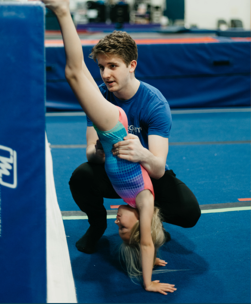 A man is helping a young girl do a handstand on a blue mat