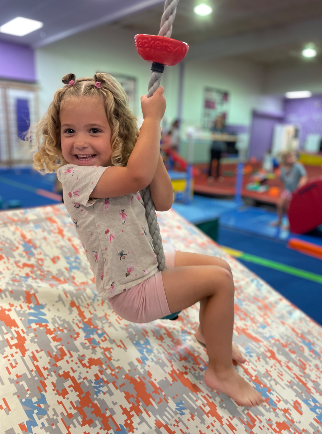 A little girl is sitting on a rope swing in a gym.