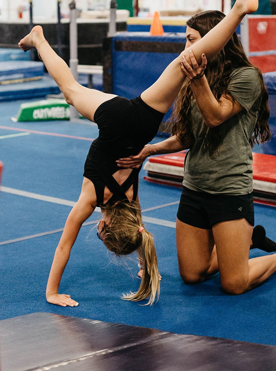 A woman is helping a young girl do a handstand in a gym.