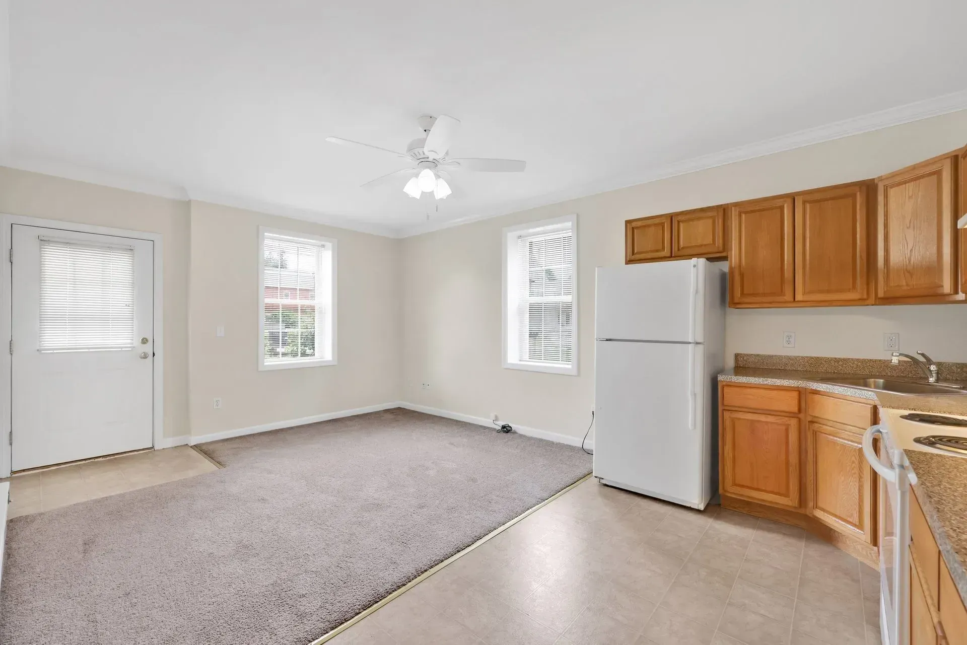 Empty kitchen and dining area with white walls, beige carpet, wood cabinets, and white appliances.