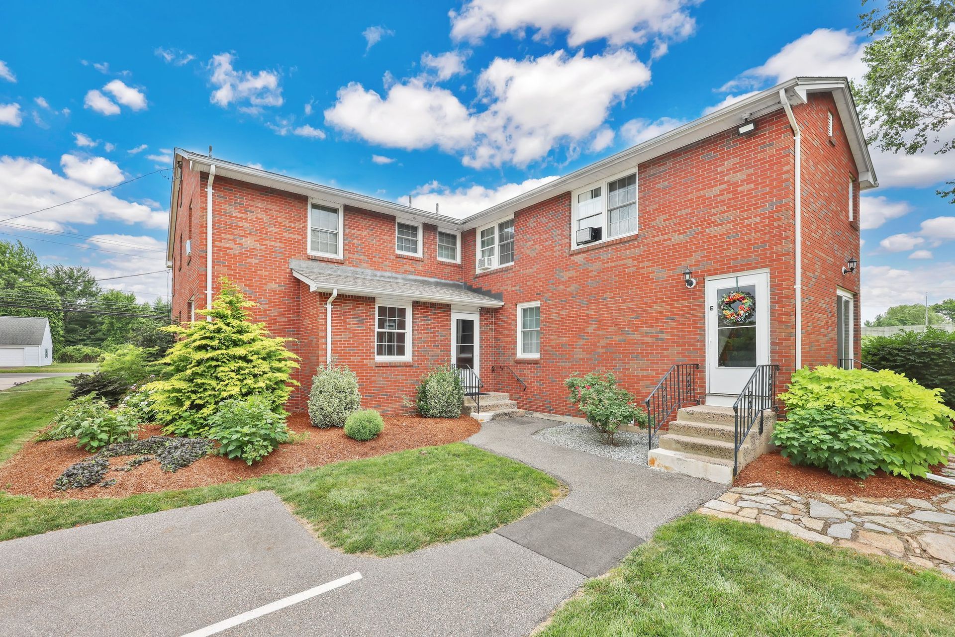 Brick townhouse exterior with front steps, walkway, shrubs, and blue sky