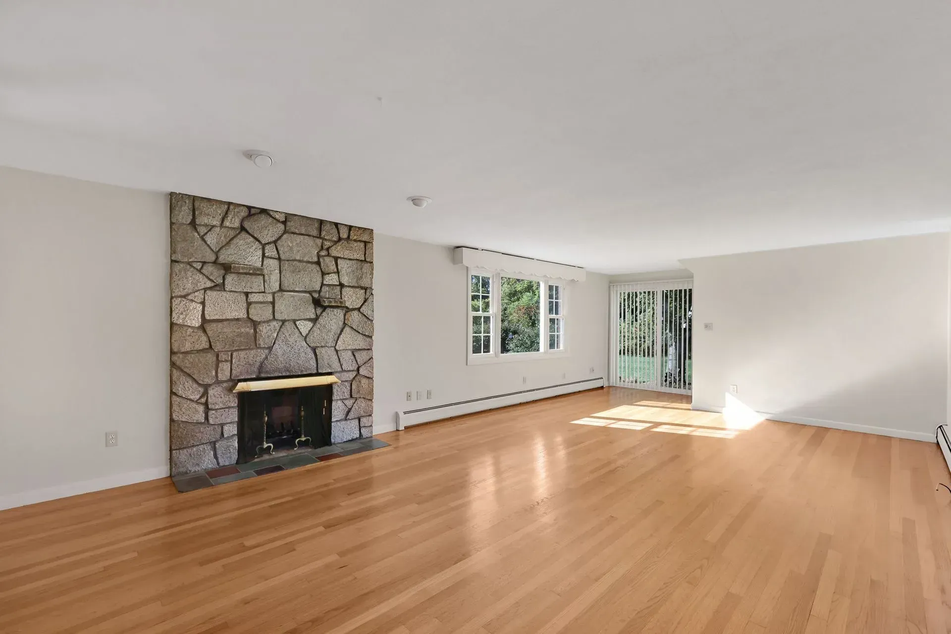 Empty living room with hardwood floors, stone fireplace, and large windows letting in daylight