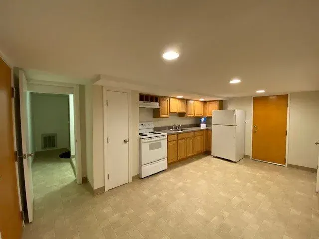 Empty apartment living area with kitchen, white appliances, wood cabinets, beige carpet, and overhead lights
