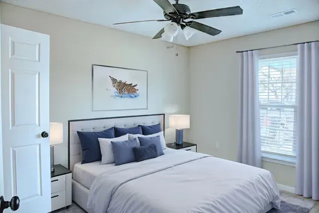 Bright bedroom with a neatly made bed, ceiling fan, nightstand lamp, and window with white curtains