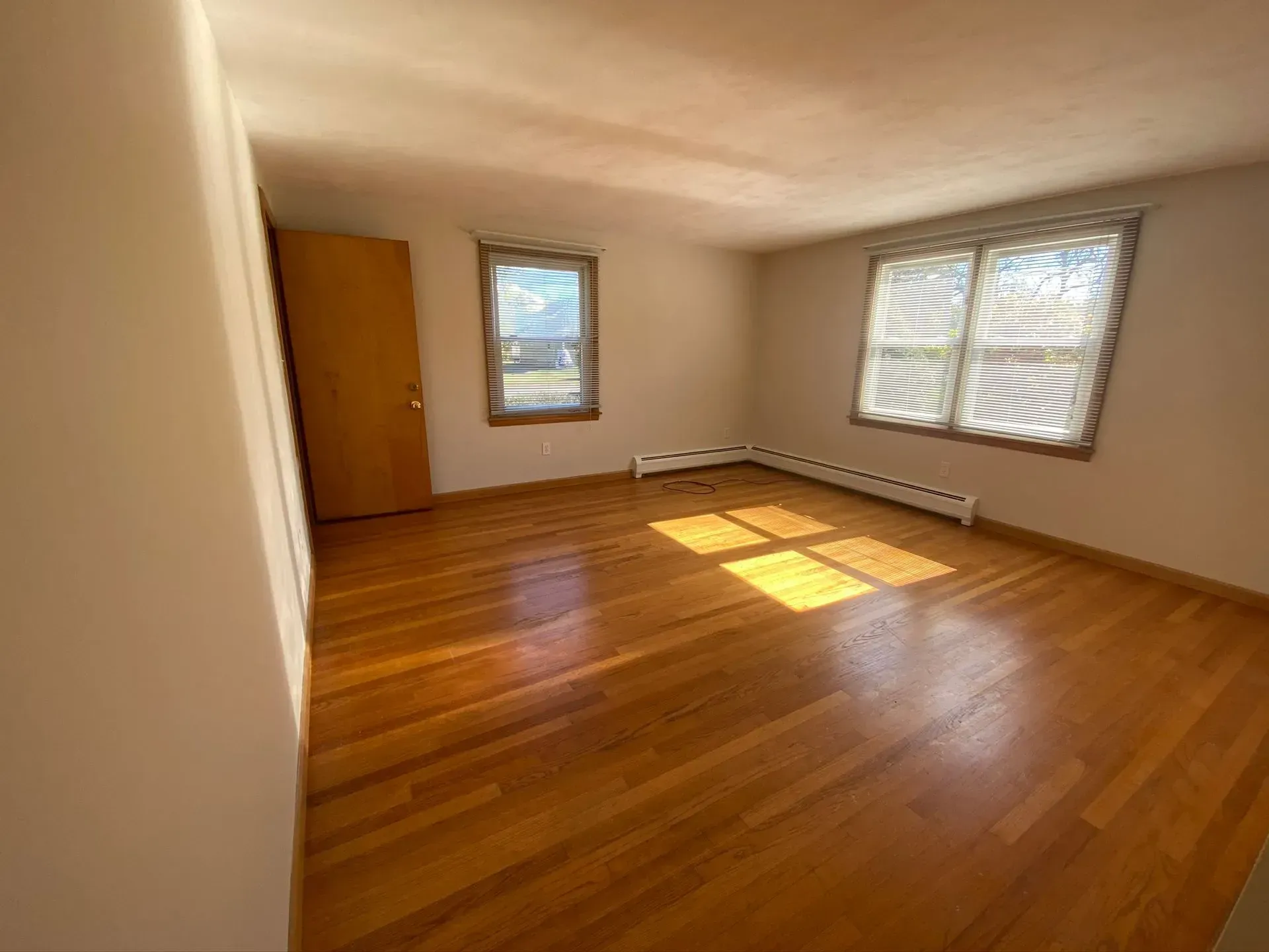 Empty room with hardwood floors, two windows, a wooden door, and sunlight on the floor.