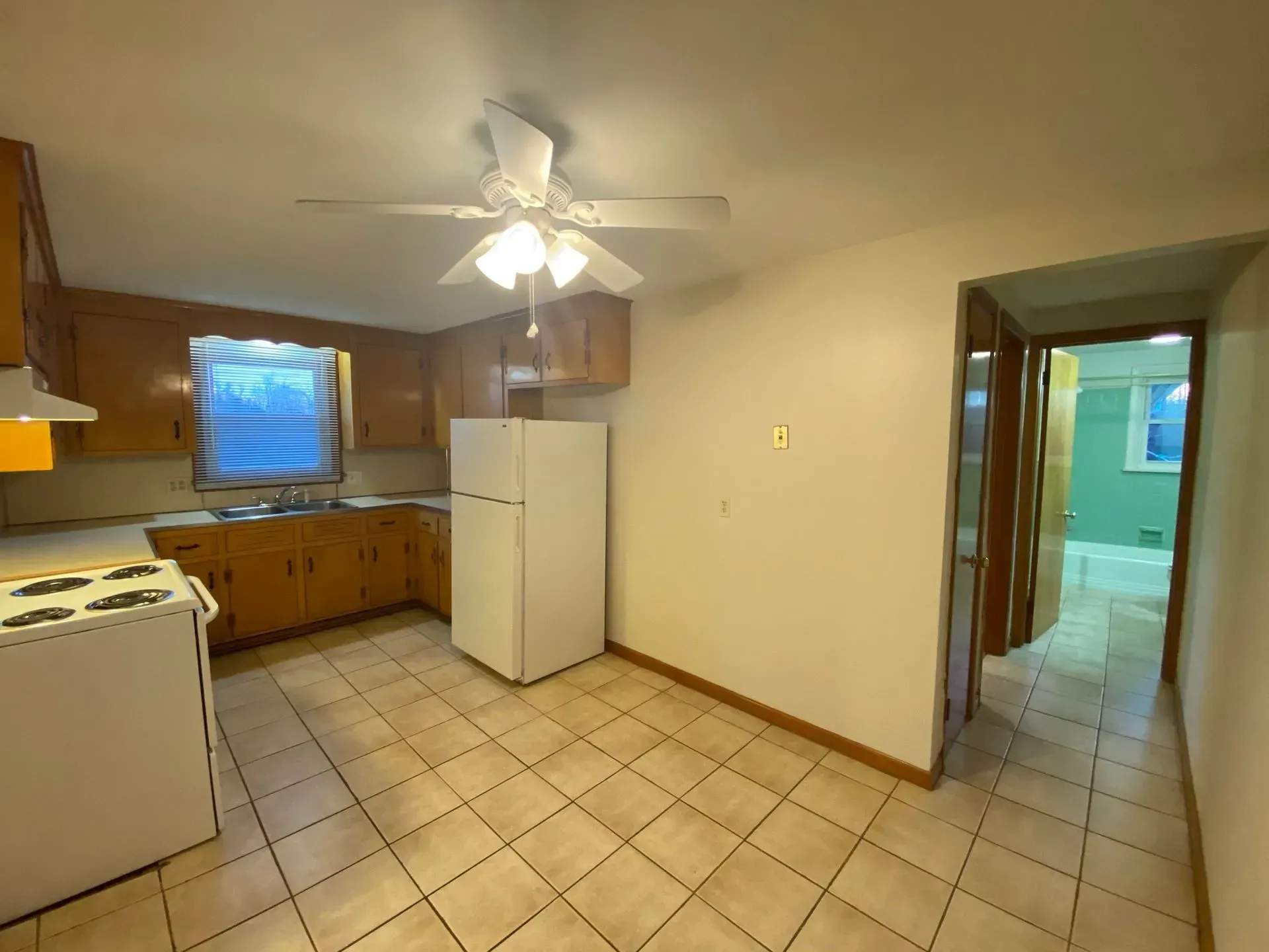 Empty kitchen with white appliances, wood cabinets, tile floor, and a doorway to a bright room