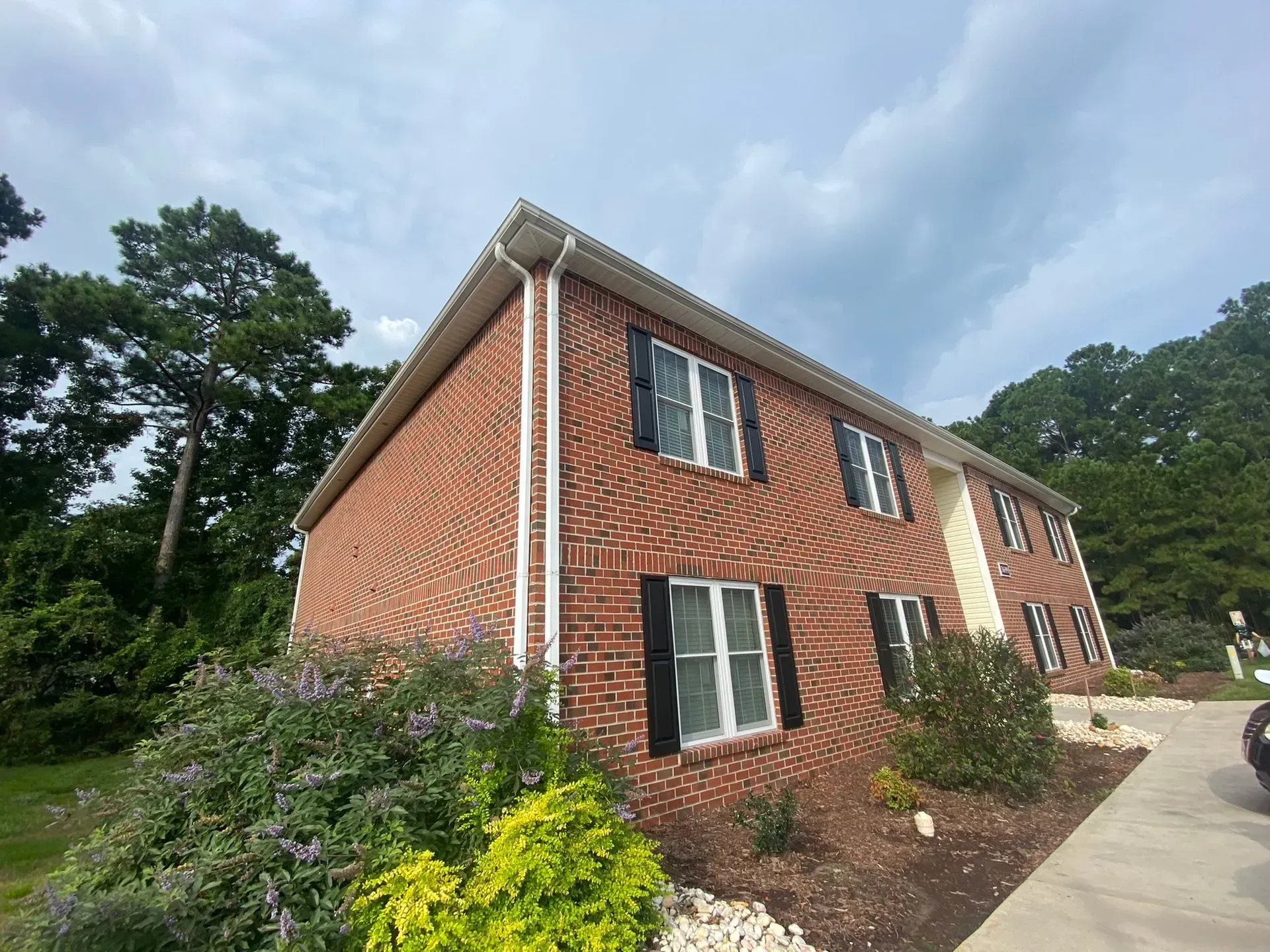 Brick apartment building with black shutters beside a paved driveway and trees