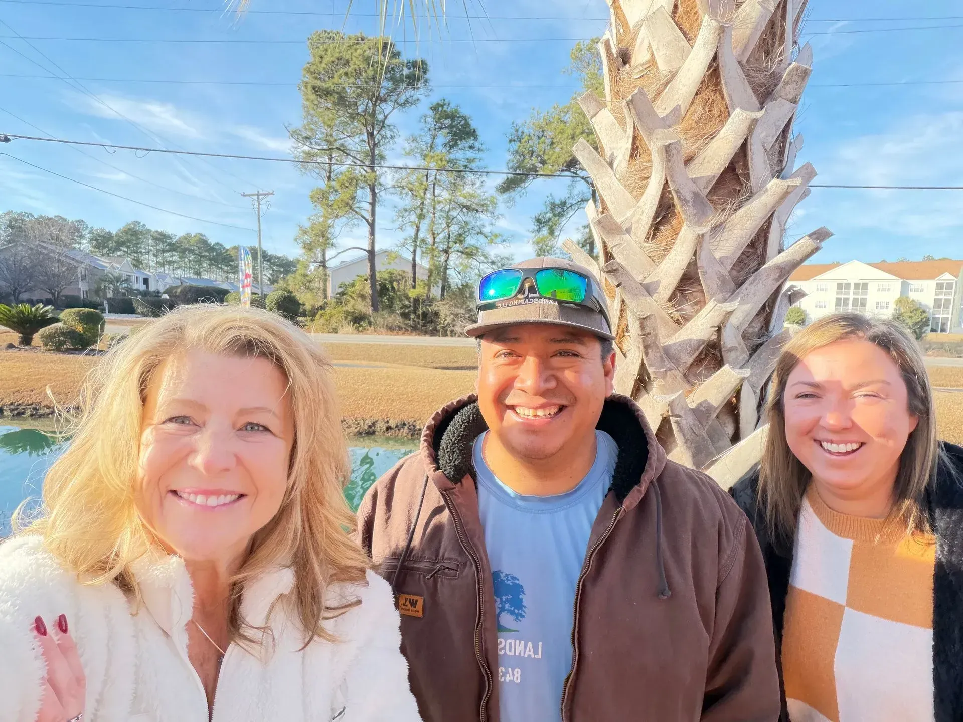 Three smiling people standing outdoors by a palm tree and pond on a sunny day